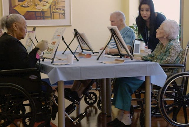 Cuatro adultos mayores pintando sobre atriles en una mesa con un miembro del personal