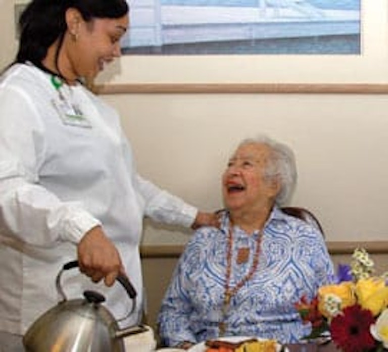 Una cuidadora de MJHS contenta con una residente mayor al lado de una tetera