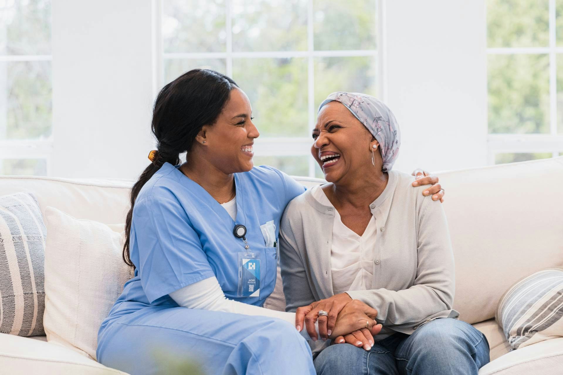 A nurse and a patient sit together on a couch, smiling at each other. The nurse has her arm around the patient