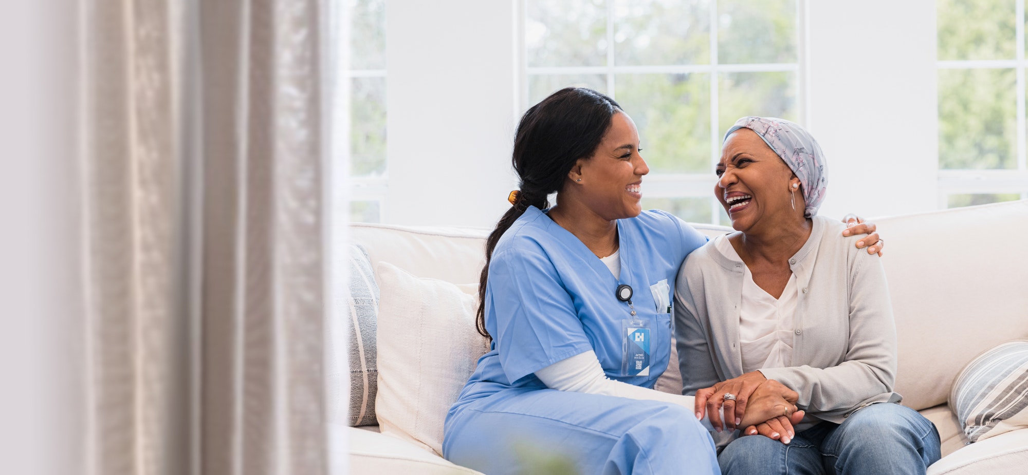 Una enfermera y un paciente se sientan juntos en un sofá y sonríen. La enfermera tiene un brazo sobre el paciente.