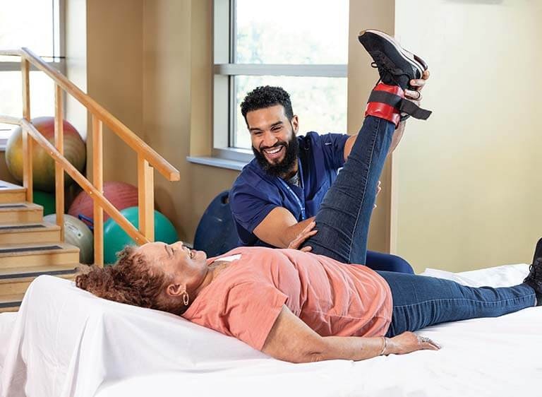 A woman laughs with her physical therapist while she performs leg stretches.