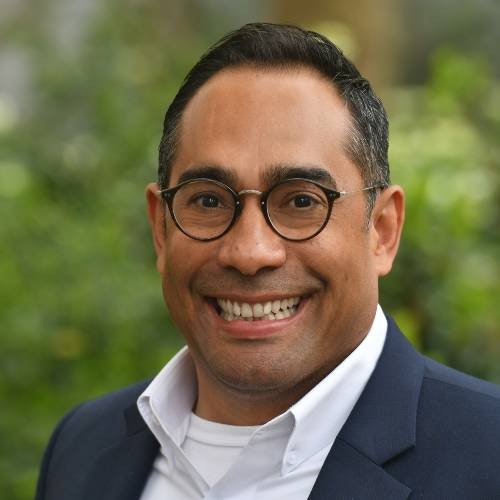 A man with short dark hair and glasses wearing a gray jacket and white collared shirt