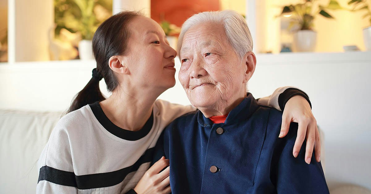 A young East Asian woman gently kisses an older East Asian woman's cheek while wrapping her arms around her shoulders in a warm, affectionate embrace.